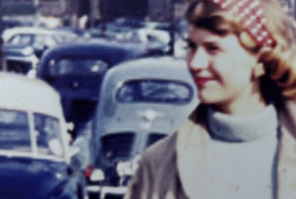 Vintage photograph of a smiling young woman in a headscarf, standing near a street filled with classic cars. Her blurred expression contrasts with the bustling background, echoing the theme of dissociation in Sylvia Plath's poems explored in 'Mad Girl’s Love Song.
