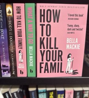 A photo of a bookshelf displaying several books, including multiple editions of How to Kill Your Family by Bella Mackie. The book features a bold pink cover with black text and an illustration of a woman holding a shovel, suggesting dark humour and crime themes. This novel explores Antisocial Personality Disorder (ASPD) through the protagonist, Grace, who calmly plans and executes murders without remorse.